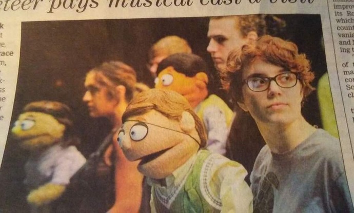 an image in a newspaper of Grayson, a white person with short, reddish-brown hair and glasses, wearing a gray-ish blue t-shirt, looking off to the side, and holding a puppet with peach-colored skin, short, brown hair, a yellowish-white button up shirt, and a green sweater vest. Behind Grayson is a line of a couple other people also holding puppets in front of a black background.