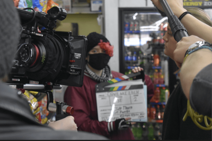 A blurry photo of Grayson, a white person with pink hair in a black beanie, a black mask, and a maroon jacket on, holding a film production slate in a convenience store. A camera, pointed at them, is in focus in the foreground