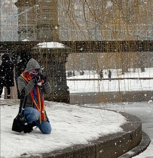 Grayson, a white person with split dyed purple and turquoise hair kneels in the snow at the edge of the pond in the Boston Public Garden, with a footbridge behind them. They are bundled up in a gray jacket, blue jeans, black boots, a purple mask, and a big rainbow scarf, and they are holding a camera and pointing it towards the pond.