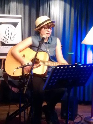 Grayson, a white person with glasses and short brown hair that sticks out of their cream-colored fedora hat that has a black ribbon around it. They are wearing a blue button up sleeveless shirt, black pants that are partially obsured in the photo, and a silver key necklace. They are sitting on a stool, holiding a guitar, and singing in front of a microphone and music stand. There is a Zing Productions logo on a stand in the background, and blue curtains.