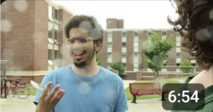 a man with a short afro, glasses, and short facial hair, wearing a blue t-shirt, holds a plastic spoon and looks down at it, confused. He is outside of a college dorm building, and a person with brown hair and a green shirt is partially in frame on the right edge of the picture.